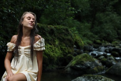 Beautiful young woman in dress sitting on rock near mountain river outdoors. Space for text