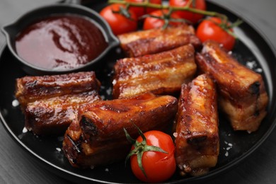 Photo of Tasty roasted pork ribs served with sauce and tomatoes on grey wooden table, closeup