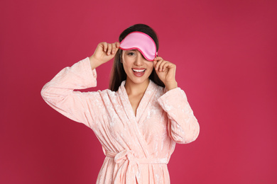 Photo of Beautiful young woman in bathrobe and eye sleeping mask on crimson background