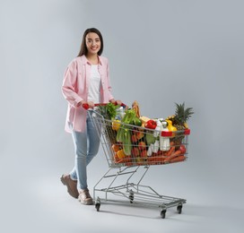 Young woman with shopping cart full of groceries on grey background