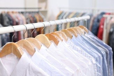 Photo of Dry-cleaning service. Many different clothes in plastic bags hanging on rack indoors, closeup