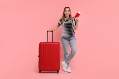 Happy young woman with passport, ticket and suitcase on pink background