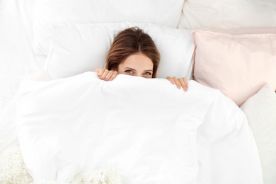 Photo of Young woman lying on soft pillows and hiding under blanket in bed at home