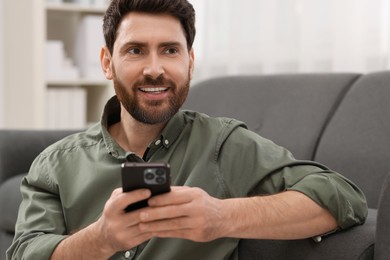 Photo of Smiling man using smartphone near sofa at home