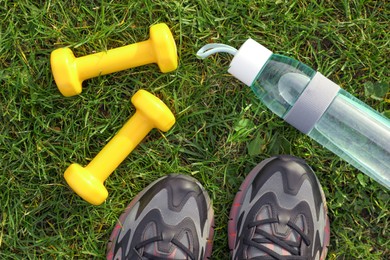 Sneakers, dumbbells and bottle of water on green grass, flat lay. Morning exercise