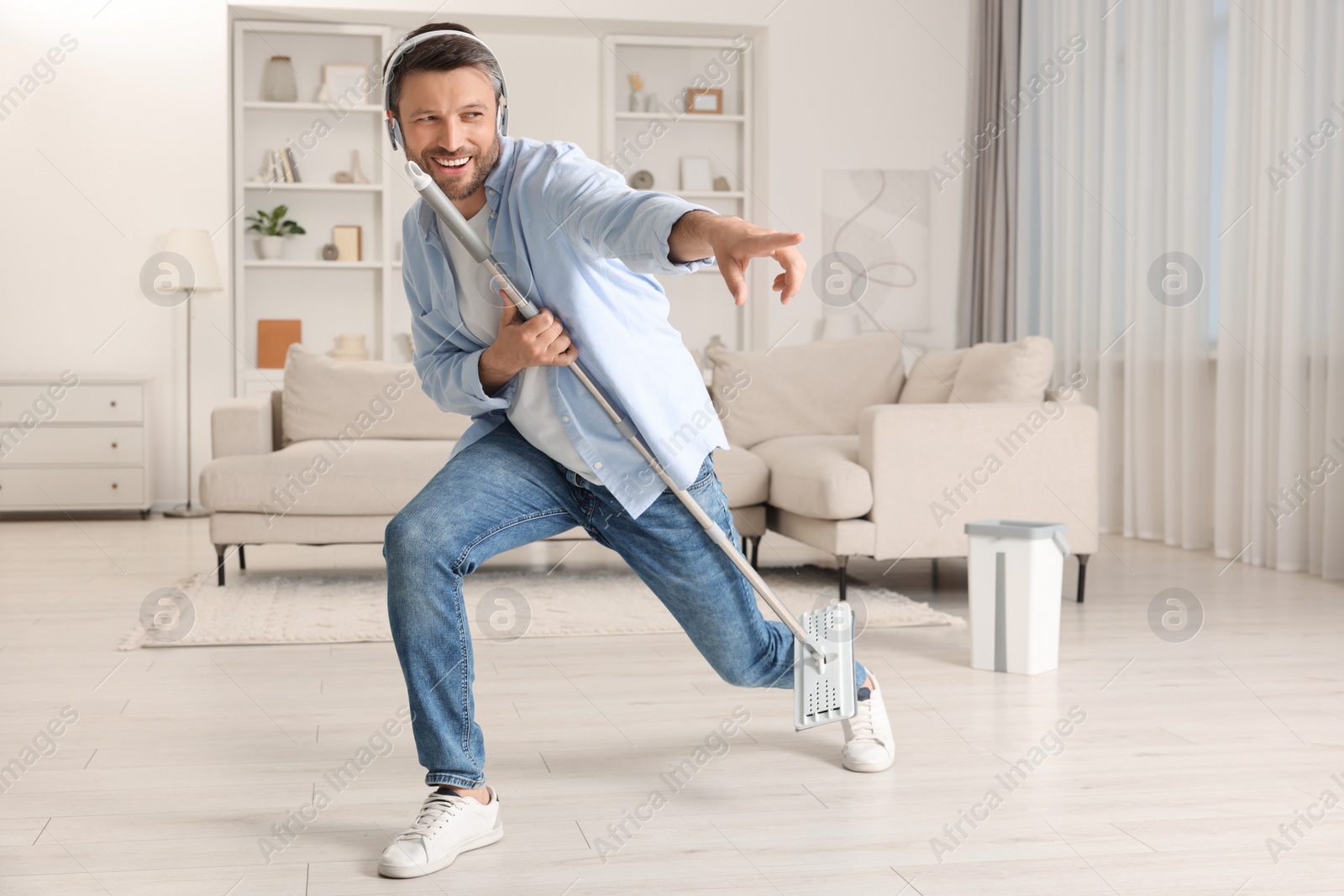 Photo of Enjoying cleaning. Happy man in headphones with mop singing while tidying up at home