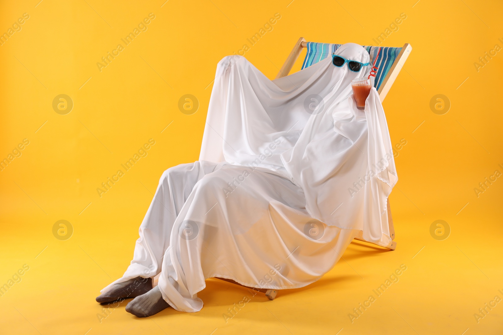 Photo of Person in ghost costume and sunglasses with glass of drink relaxing on deckchair against yellow background