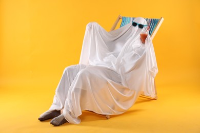 Person in ghost costume and sunglasses with glass of drink relaxing on deckchair against yellow background