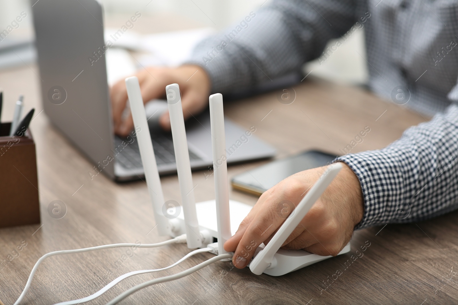 Photo of Man with laptop inserting cable into Wi-Fi router at wooden table indoors, closeup