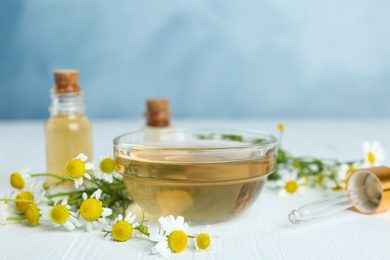 Bowl of essential oil and fresh chamomiles on white wooden table