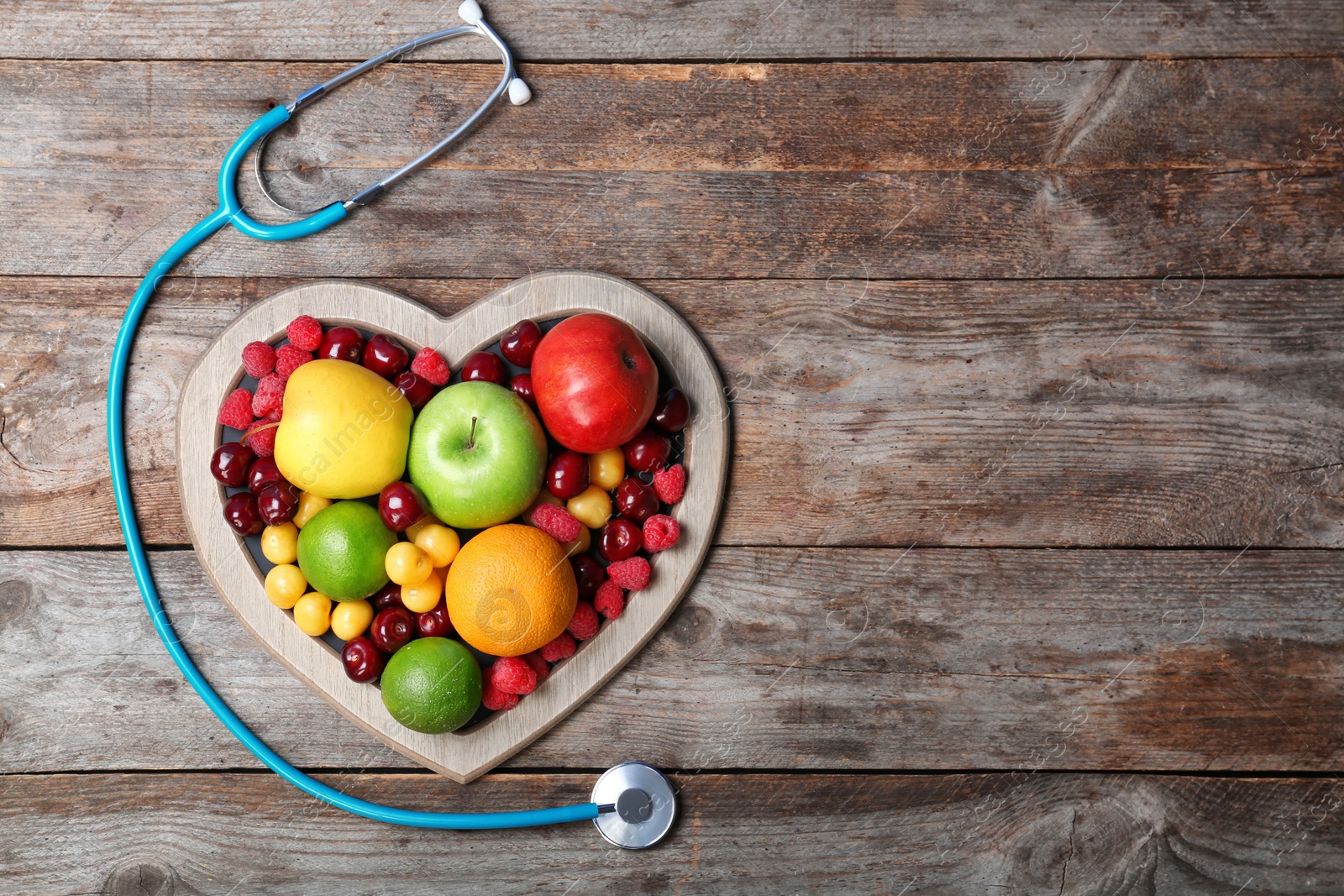 Photo of Heart shaped plate with fresh fruits and stethoscope on wooden table, top view. Cardiac diet