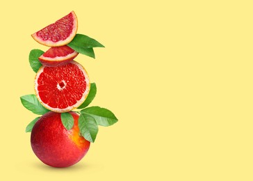 Stacked cut and whole red oranges with green leaves on pale light yellow background, space for text