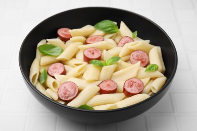 Tasty pasta with smoked sausage and basil in bowl on white tiled table, closeup