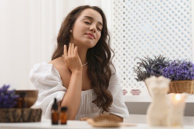 Photo of Beautiful young woman applying essential oil onto neck at table indoors