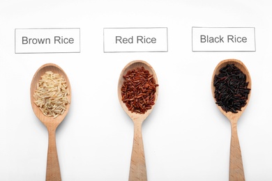 Spoons with different types of uncooked rice and cards on white background, top view