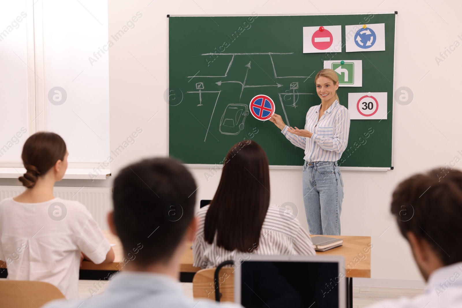 Photo of Teacher showing No Stopping road sign near chalkboard during lesson in driving school