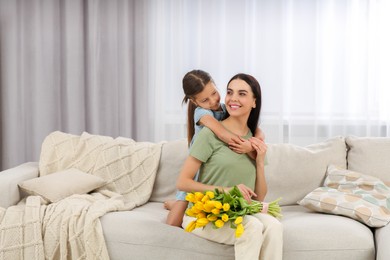 Photo of Happy woman and her cute daughter with bouquet of yellow tulips on sofa at home. Mother's day celebration