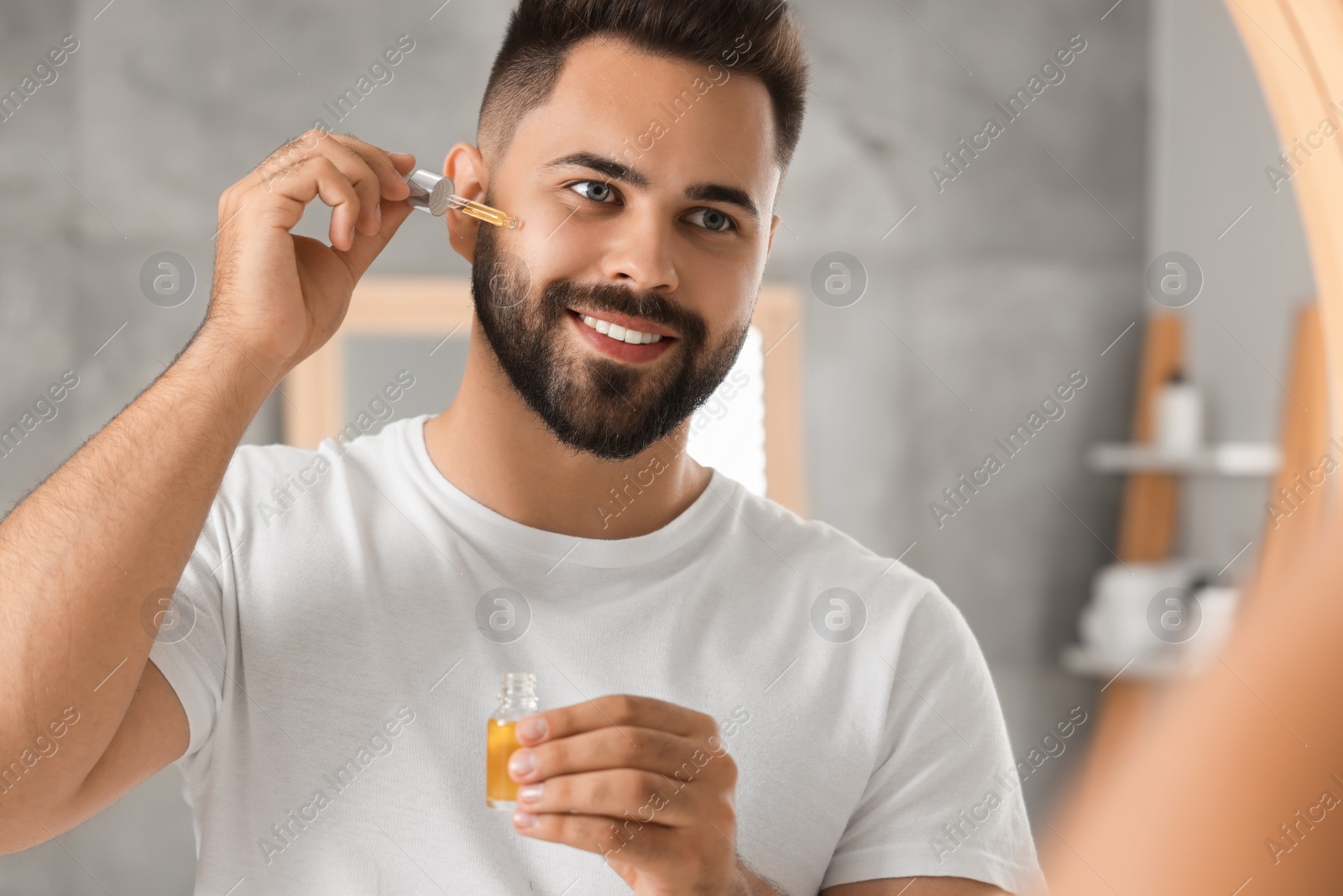 Photo of Handsome man applying cosmetic serum onto his face near mirror indoors