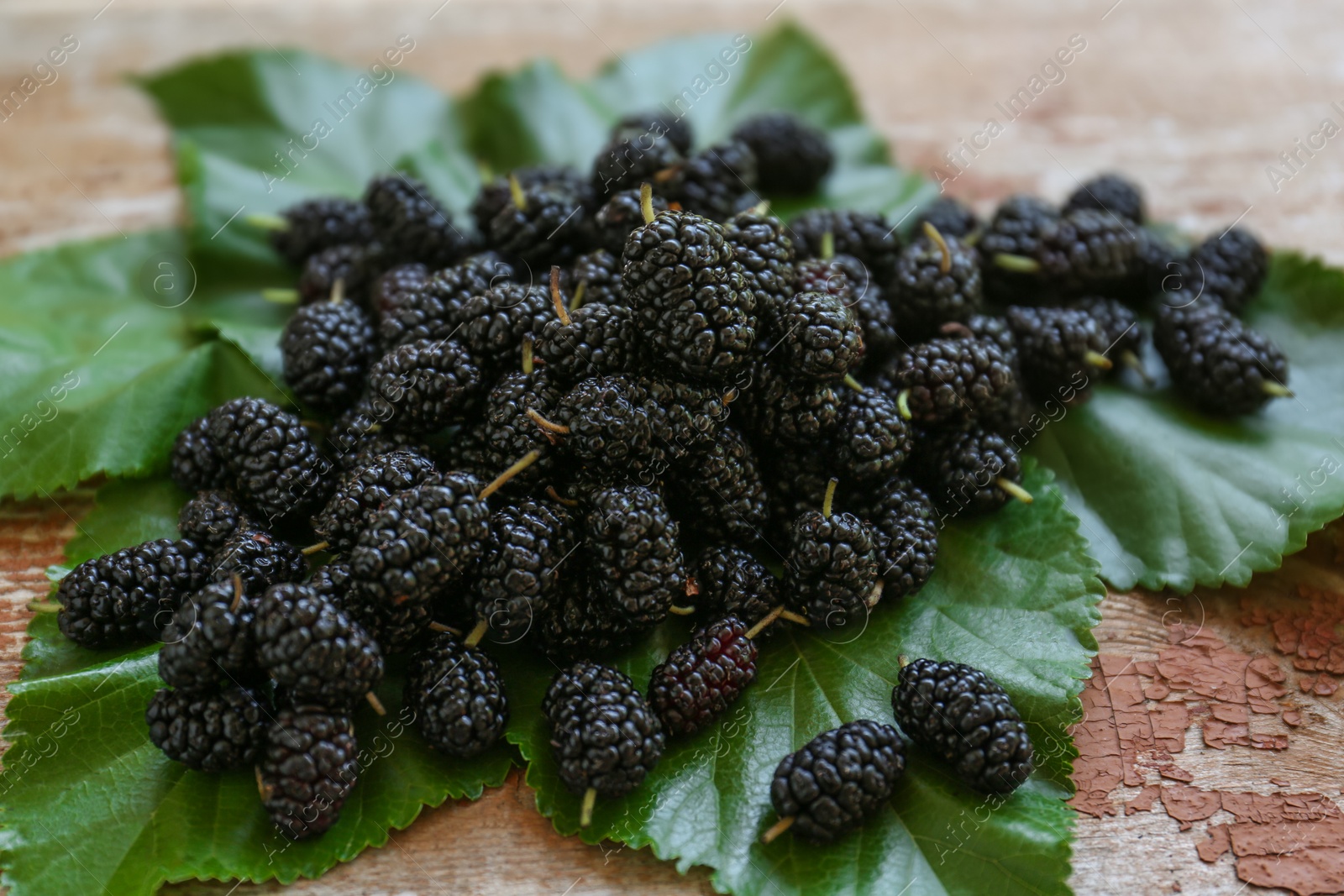 Photo of Heap of delicious ripe black mulberries and green leaves on wooden table, closeup