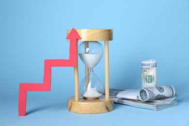 Photo of Economic profit. Hourglass, banknotes and arrow on light blue background