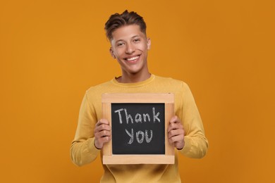 Photo of Happy man holding small chalkboard with phrase Thank You on orange background