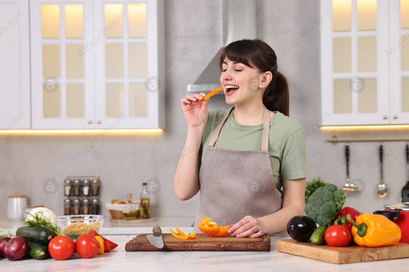 Photo of Young housewife tasting cut bell pepper while cooking at white marble table in kitchen