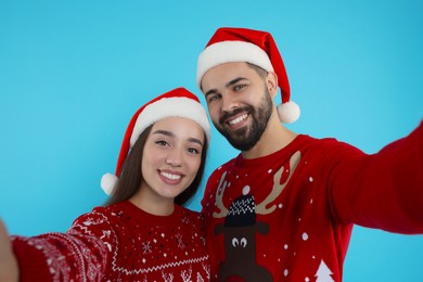 Happy young couple in Christmas sweaters and Santa hats taking selfie on light blue background