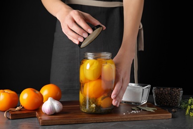 Photo of Woman opening jar with pickled tomatoes at grey table against black background, closeup