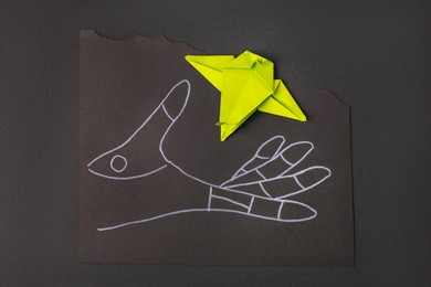 Drawn hand and green paper bird on black background, top view