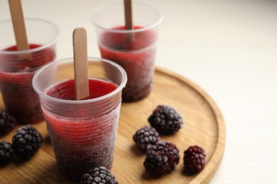 Tasty blackberry ice pops in plastic cups on white wooden table, closeup. Fruit popsicle