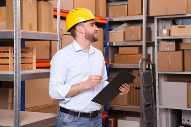 Photo of Young man with clipboard near rack of cardboard boxes at warehouse