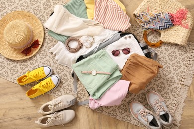 Open suitcase full of clothes and summer accessories near shoes on floor, top view