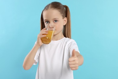 Photo of Little girl drinking fresh juice and showing thumbs up on light blue background