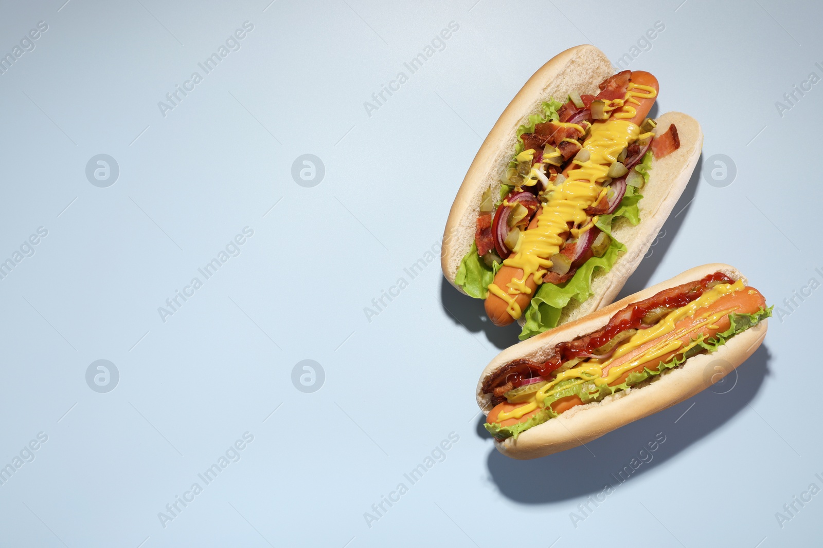 Photo of Delicious hot dogs with bacon, lettuce, onion and pickle on light blue background, flat lay. Space for text