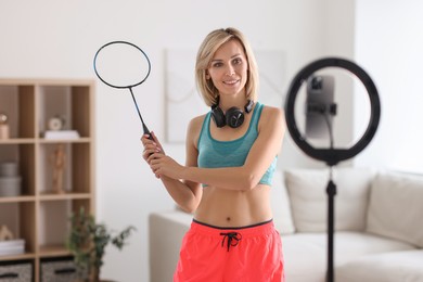 Photo of Smiling sports blogger holding badminton racket while streaming online fitness lesson with smartphone at home