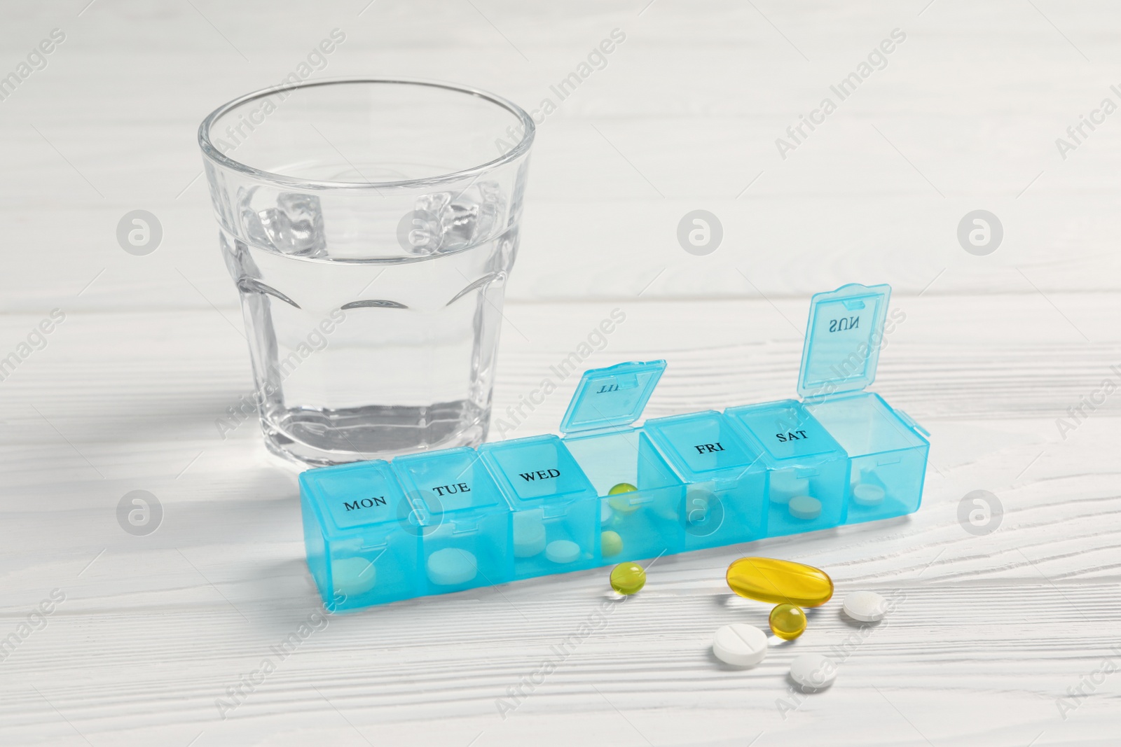 Photo of Weekly pill box with medicaments and glass of water on white wooden table