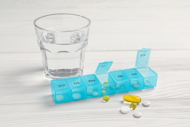 Photo of Weekly pill box with medicaments and glass of water on white wooden table