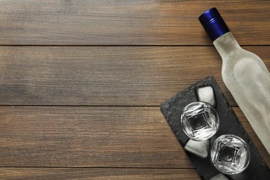 Photo of Bottle of vodka, shot glasses and ice cubes on wooden table, flat lay. Space for text