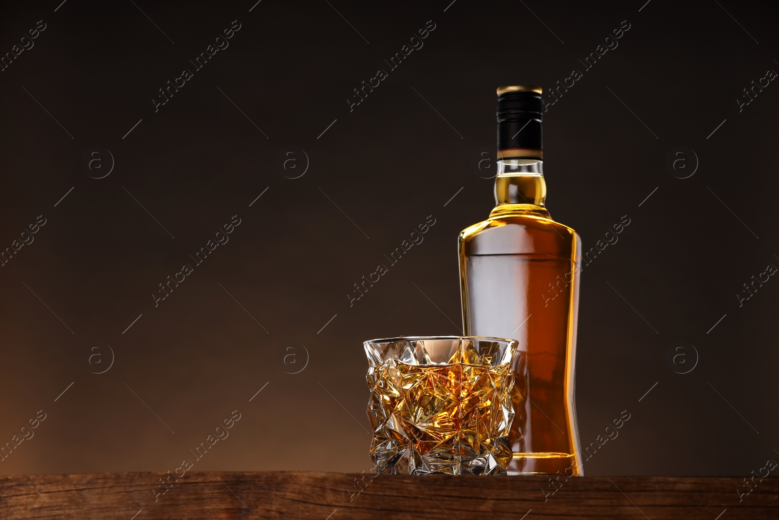 Photo of Whiskey in glass and bottle on wooden table against brown background, space for text