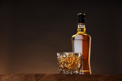 Photo of Whiskey in glass and bottle on wooden table against brown background, space for text