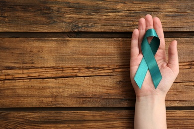 Woman holding teal awareness ribbon on wooden background, top view with space for text. Symbol of social and medical issues