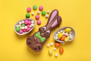 Photo of Chocolate Easter bunny, halves of egg and candies on yellow background, flat lay