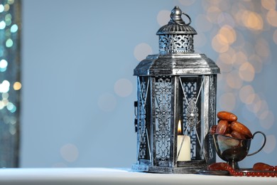 Photo of Arabic lantern, misbaha and dates on table against blurred lights, space for text