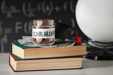 Photo of Scholarship concept. Glass jar with coins, graduation cap, dollar banknotes and books on white wooden table