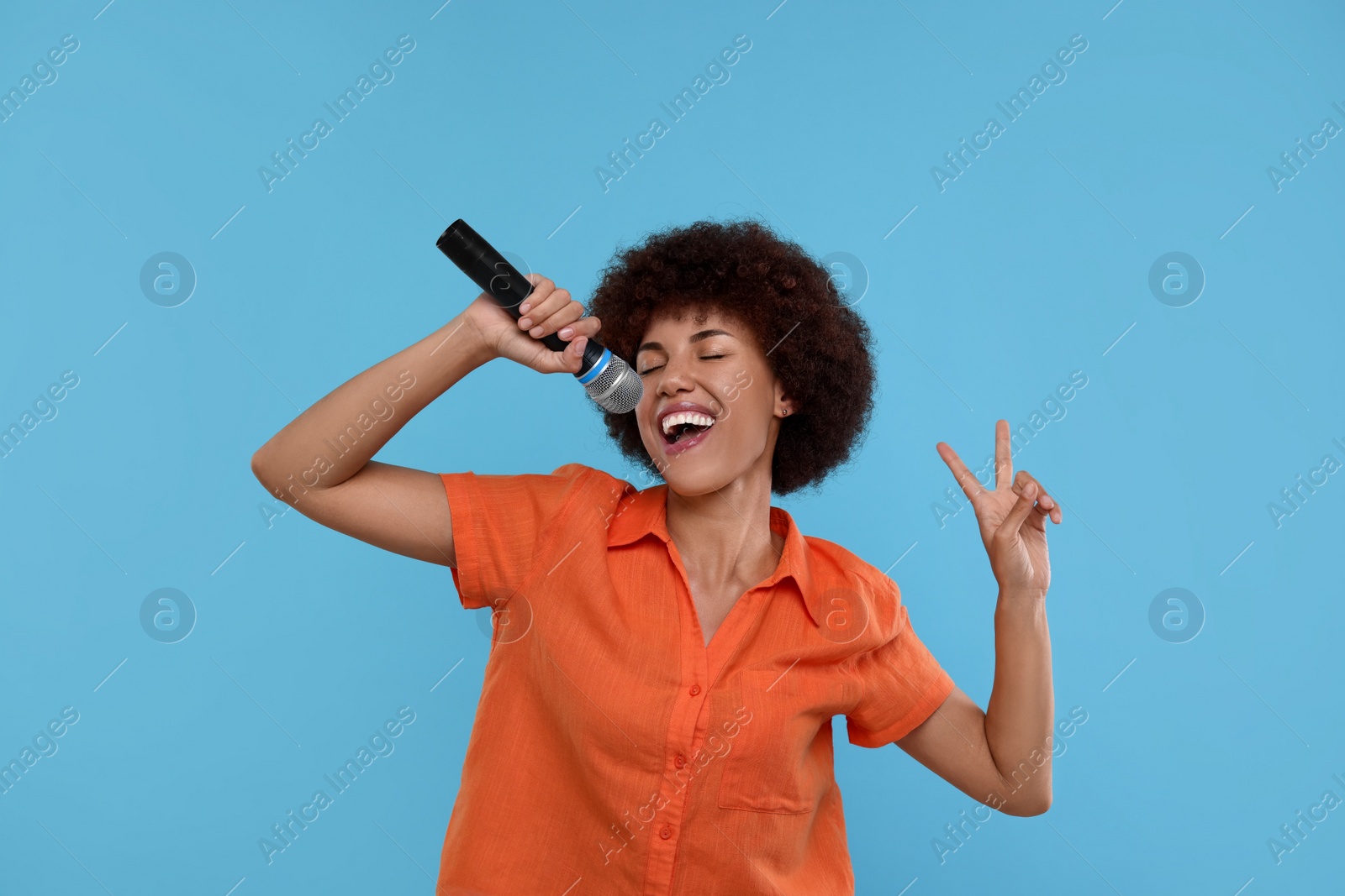 Photo of Curly young woman with microphone singing and showing victory sign on light blue background