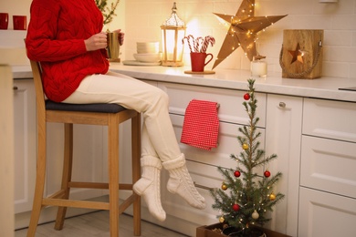 Photo of Woman with cup of hot drink sitting on bar stool in kitchen decorated for Christmas