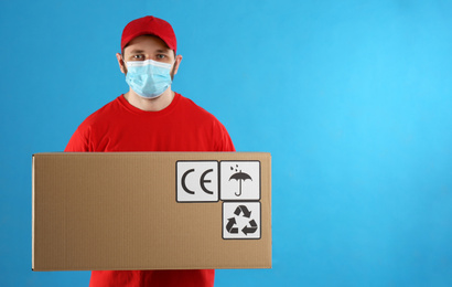 Photo of Courier in mask holding cardboard box with different packaging symbols on blue background. Parcel delivery