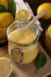 Photo of Delicious lemon curd in glass jar, spoon, fresh citrus fruits and green leaves on table
