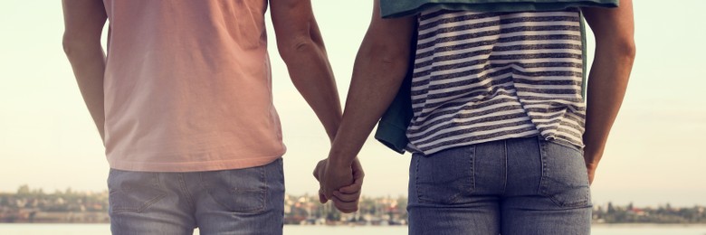 Gay couple holding hands together near river on sunny day, back view. Banner design