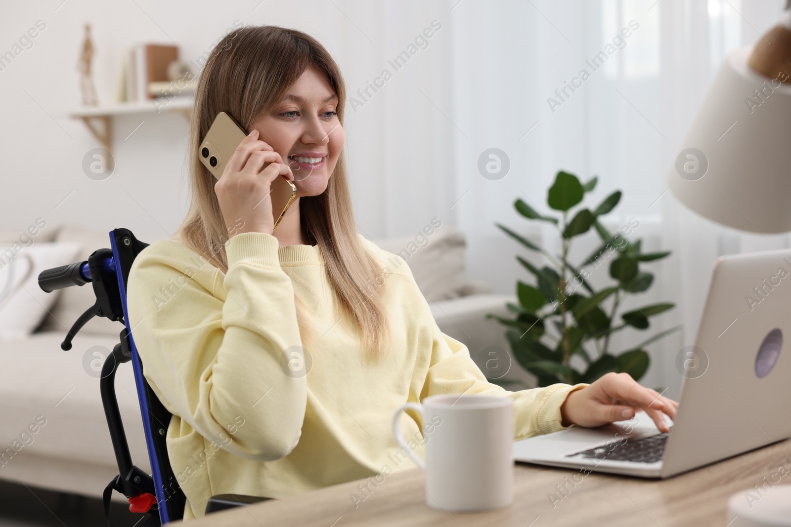 Photo of Woman in wheelchair talking on smartphone while using laptop at table in home office